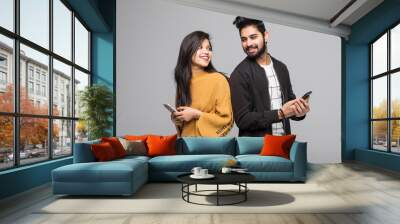 Beautiful young indian couple holding mobile phones and standing back to back against grey background Wall mural